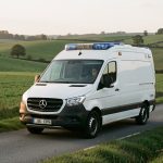 Ambulance blanche roulant sur une route rurale du Gers près d’Eauze, au milieu d’un paysage vallonné baigné d’une lumière naturelle réaliste.